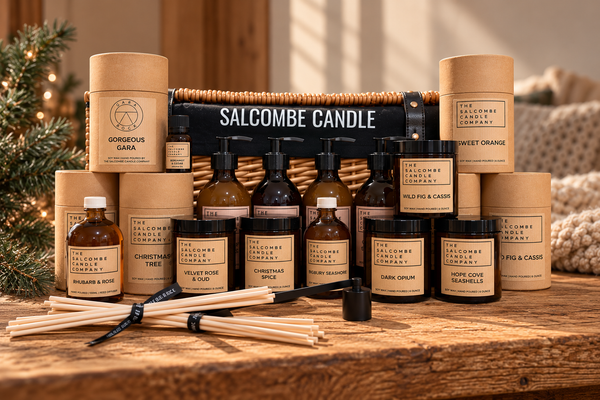 A Salcombe Candle gift set arranged on a rustic wooden table, featuring amber glass bottles, cylindrical kraft paper tubes, and a wicker basket with a black Salcombe Candle sign. A black reed diffuser stick and wooden reeds are placed in front, with holiday greenery and a cozy knit blanket softly blurred in the background.