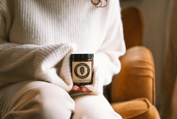 A person in a cozy white ribbed sweater holds a small jar candle labeled 'Pomegranate & Rosemary' in their lap while sitting in a warm brown armchair.