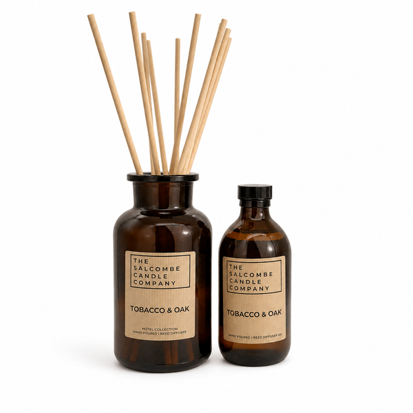 The Salcombe Candle Company Tobacco & Oak reed diffuser set, featuring an amber glass bottle with natural wood reeds and a matching amber glass bottle with a black cap, both with kraft paper labels, on a white background.