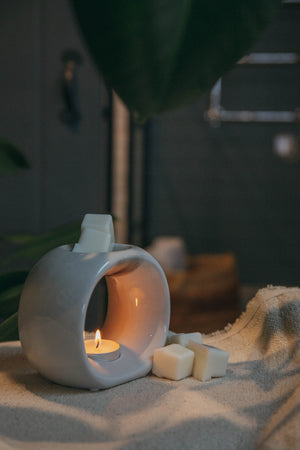 White ceramic burner with a lit candle on a towel, surrounded by wax melt cubes.